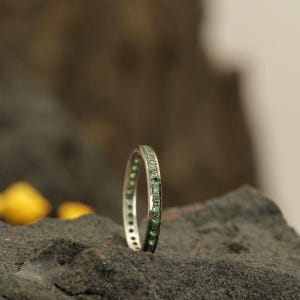 May include: A thin silver-toned metal band ring featuring a continuous row of small, round, vibrant green gemstones set along the outer circumference. The ring is positioned upright, showcasing the uniform placement of the green stones.