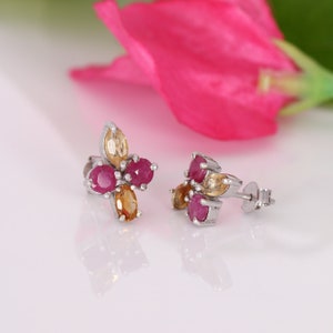 May include: A pair of silver stud earrings with a flower design. The earrings feature red and yellow gemstones.