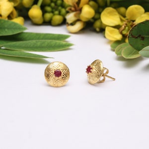 Op de afbeelding: Een paar gouden oorbelletjes met een rode edelsteen in het midden van elke oorbel. De oorbelletjes hebben een geometrisch ontwerp.