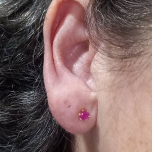 Op de afbeelding: Een close-up van iemands oor met een kleine roze edelsteen oorring. De oorring heeft een gouden zetting met een kleine, decoratieve rand.