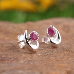 Può includere: Un paio di orecchini a lobo in argento sterling con una gemma di rubino rotonda, sfaccettata e di colore rosa intenso, incastonata in una montatura in metallo liscia e curva a forma di mezzaluna, con chiusure a perno standard.