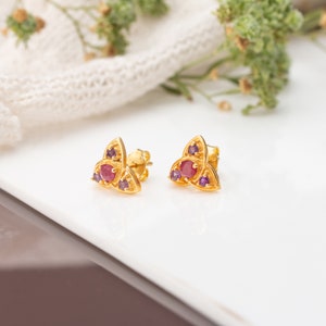 Könnte beinhalten: Ein Paar goldene Ohrringe mit einem keltischen Knotendesign. Jeder Ohrring hat einen roten Rubin und zwei violette Amethyste.