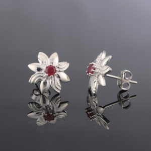Op de afbeelding: Een paar zilveren oorbellen met rode edelsteenbloem accenten. De oorbellen hebben een stervormig ontwerp met een kleine rode edelsteen in het midden van elke bloem.