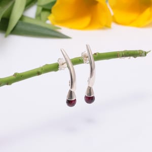 Może przedstawiać: Para srebrnych kolczyków z zakrzywionym wzorem i wiszącym czerwonym kamieniem szlachetnym.