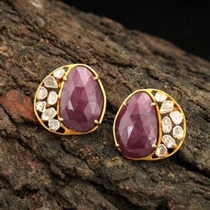 Puede incluir: Un par de pendientes de botón dorados. Cada pendiente presenta una gema grande, facetada, de forma ovalada, de color rojo intenso, acentuada con un grupo de gemas más pequeñas y transparentes. Los pendientes están colocados sobre una superficie de madera oscura y texturizada.