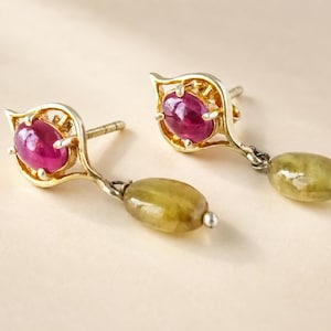 Può includere: Un paio di orecchini color oro con una gemma centrale ovale color rosa intenso taglio cabochon incastonata in una montatura di metallo a punta, con una gemma ovale liscia color verde oliva appesa alla base di ogni perno.