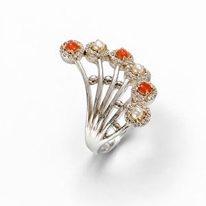 Puede incluir: Un anillo de plata con un diseño en forma de abanico con seis puntas radiantes. Cada punta está rematada con un engaste circular que contiene un cabujón de coral naranja o una perla blanca, rodeado por un halo de pequeñas piedras transparentes.