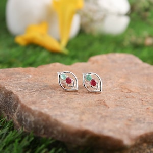 Könnte beinhalten: Ein Paar silberne Ohrringe mit einem Blattdesign. Jeder Ohrring hat einen roten und grünen Edelstein.