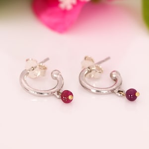 May include: A pair of silver hoop earrings with a small, dangling red bead and a gold accent. The earrings have a simple, elegant design, perfect for everyday wear. The earrings are displayed on a white surface with a blurred pink background.