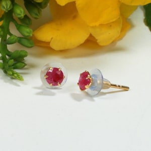 May include: A pair of stud earrings featuring round, faceted, deep pink gemstones set in gold-toned metal prongs. Each earring includes a clear, circular plastic backing and a gold-toned post, showcasing a simple and classic design for everyday wear.