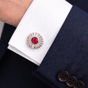 Pode incluir: Close-up de uma abotoadura prateada com uma pedra preciosa vermelha no centro, contra uma manga de camisa branca e um fato azul marinho. A abotoadura tem um design radial.