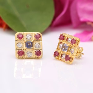 Op de afbeelding: Gouden vierkante oorbel studs met een blauwe edelsteen in het midden, omringd door kleinere rode en witte edelstenen.