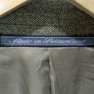 May include: Close-up of a brown and green tweed fabric with a black label that reads "Made in Switzerland".