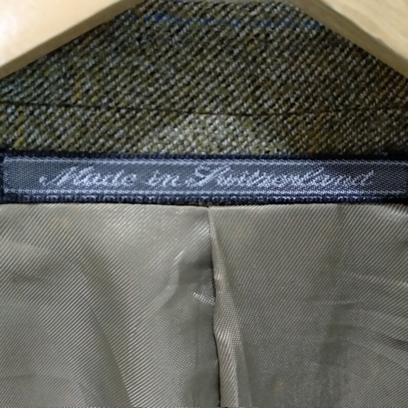 May include: Close-up of a brown and green tweed fabric with a black label that reads "Made in Switzerland".