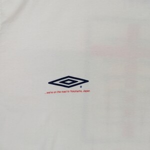 May include: White t-shirt with a navy blue diamond logo and the text "...we're on the road to Yokohama, Japan" in red.