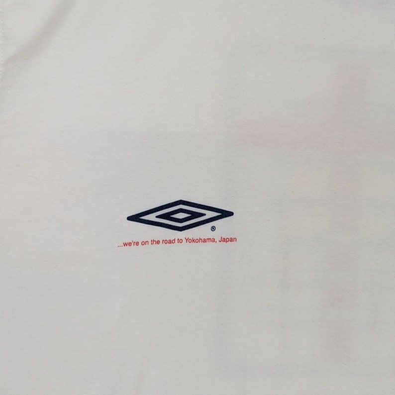 May include: White t-shirt with a navy blue diamond logo and the text "...we're on the road to Yokohama, Japan" in red.