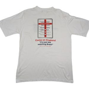 May include: White t-shirt with a graphic of a scorecard showing England's wins in the 1998 World Cup. The text "Come on England" and "It's just like watching Brazil" is printed below the scorecard.
