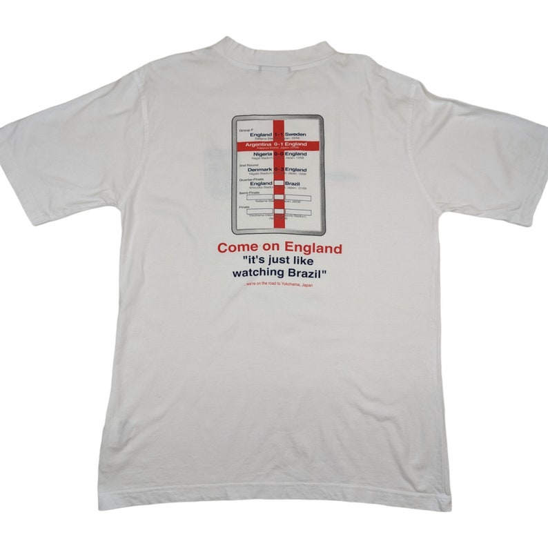 May include: White t-shirt with a graphic of a scorecard showing England's wins in the 1998 World Cup. The text "Come on England" and "It's just like watching Brazil" is printed below the scorecard.
