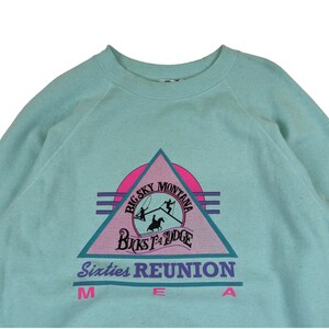 Puede incluir: Una sudadera de color azul claro con un estampado gr&aacute;fico rosa, azul y negro de un tri&aacute;ngulo con el texto "Big Sky Montana" y "Bucks T-4 Lodge" en su interior. El texto "Sixties Reunion" est&aacute; impreso debajo del tri&aacute;ngulo en morado y negro.