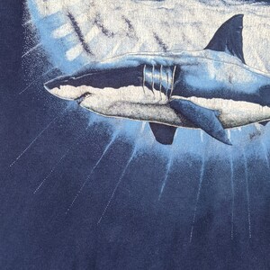 Peut inclure: Un grand requin blanc nageant dans l'eau bleue. Le requin est repr&eacute;sent&eacute; dans un style r&eacute;aliste avec des &eacute;cailles et des nageoires d&eacute;taill&eacute;es. L'arri&egrave;re-plan est bleu fonc&eacute; avec des stries bleu clair, sugg&eacute;rant la surface de l'eau.