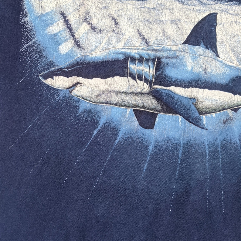 Peut inclure: Un grand requin blanc nageant dans l'eau bleue. Le requin est repr&eacute;sent&eacute; dans un style r&eacute;aliste avec des &eacute;cailles et des nageoires d&eacute;taill&eacute;es. L'arri&egrave;re-plan est bleu fonc&eacute; avec des stries bleu clair, sugg&eacute;rant la surface de l'eau.