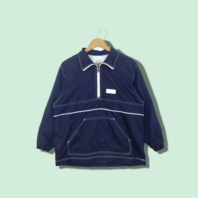May include: A navy blue windbreaker jacket with white stitching and a white zipper. The jacket has a small white patch on the chest with the word "OG" on it.