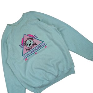 Puede incluir: Una sudadera azul claro con un estampado gr&aacute;fico rosa, morado y azul de un tri&aacute;ngulo con el texto "Big Sky Montana, Bust'n'Ridge, Sixties Reunion Area".