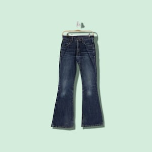 Puede incluir: Un par de pantalones vaqueros de campana de mezclilla azul oscuro colgados de una percha de madera sobre un fondo verde claro.