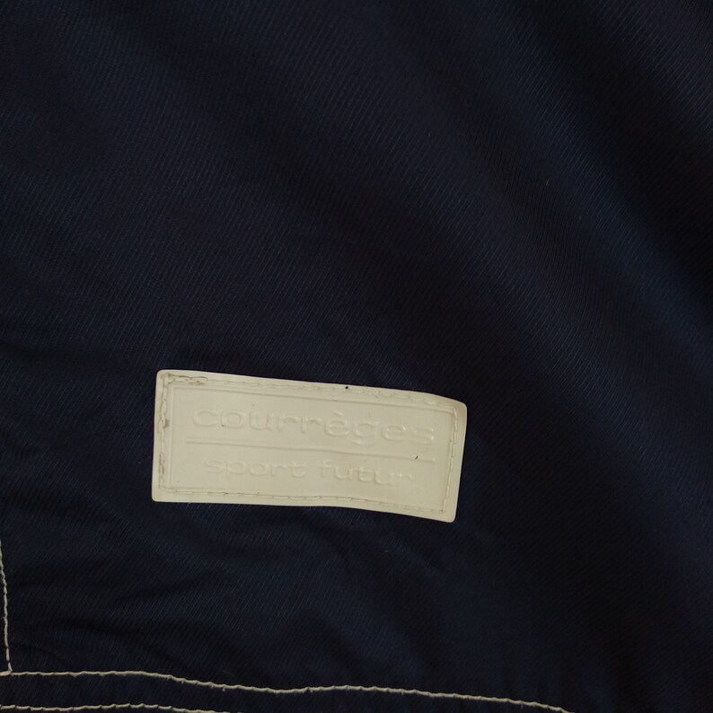 May include: A white label with the text "Courr&egrave;ges Sport futur" on a dark blue fabric background.
