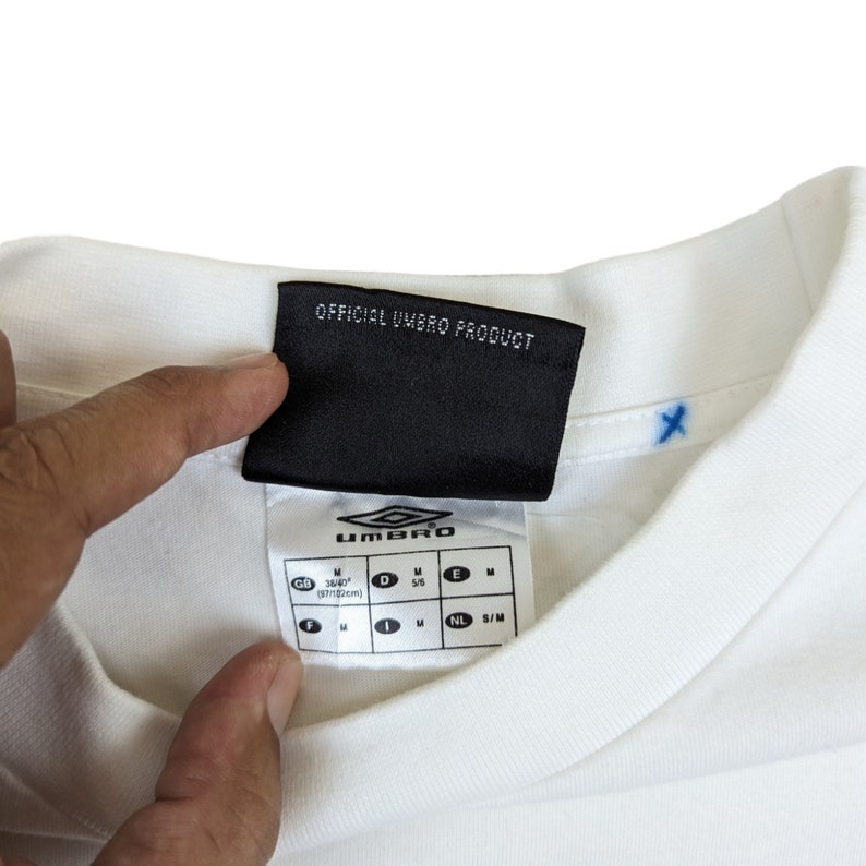 May include: White t-shirt with a black tag that says "OFFICIAL UMBRO PRODUCT" and a size chart with measurements in inches and centimeters.