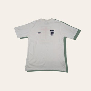 May include: White short-sleeved t-shirt with a blue and red England crest and a white and blue Umbro logo. The shirt has the text "England" above the crest and "Football Association" below the crest.