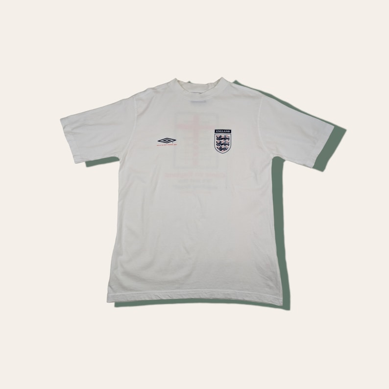 May include: White short-sleeved t-shirt with a blue and red England crest and a white and blue Umbro logo. The shirt has the text "England" above the crest and "Football Association" below the crest.