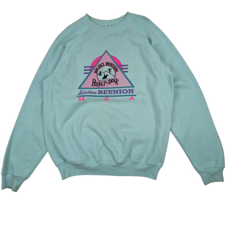 Puede incluir: Una sudadera de color azul claro con un estampado gr&aacute;fico rosa, morado y blanco de un tri&aacute;ngulo con el texto "Big Sky Montana" y "Big Sky Lodge" dentro del tri&aacute;ngulo. El texto "Sixties Reunion" est&aacute; debajo del tri&aacute;ngulo con el texto "MEA" debajo.