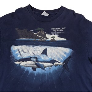 Peut inclure: Un t-shirt bleu marine avec un graphique d'un grand requin blanc nageant dans l'oc&eacute;an. Le texte "Monterey Bay Aquarium" est imprim&eacute; sur le t-shirt. Des aimants puissants et la recherche, sauvons les oc&eacute;ans le long de la c&ocirc;te californienne.
