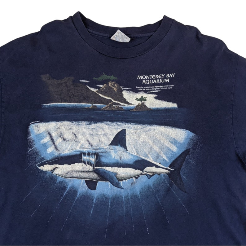 Peut inclure: Un t-shirt bleu marine avec un graphique d'un grand requin blanc nageant dans l'oc&eacute;an. Le texte "Monterey Bay Aquarium" est imprim&eacute; sur le t-shirt. Des aimants puissants et la recherche, sauvons les oc&eacute;ans le long de la c&ocirc;te californienne.