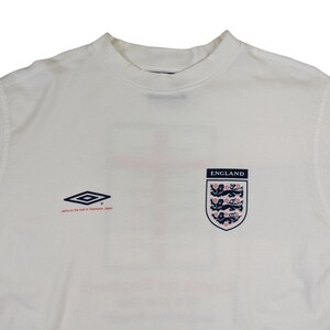 May include: White t-shirt with a navy blue England crest and the text "ENGLAND" above it. The shirt also features a navy blue and white diamond logo and the text "we're on the road to Yokohama, Japan".