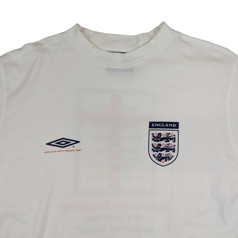 May include: White t-shirt with a navy blue England crest and the text "ENGLAND" above it. The shirt also features a navy blue and white diamond logo and the text "we're on the road to Yokohama, Japan".