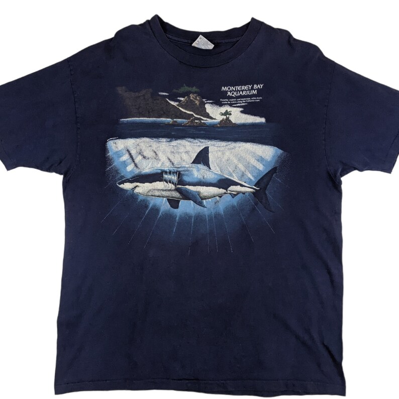 Peut inclure: Un t-shirt bleu marine avec un motif repr&eacute;sentant un grand requin blanc nageant dans l'oc&eacute;an. Le motif comprend &eacute;galement une petite &icirc;le avec des arbres et le texte "Monterey Bay Aquarium".
