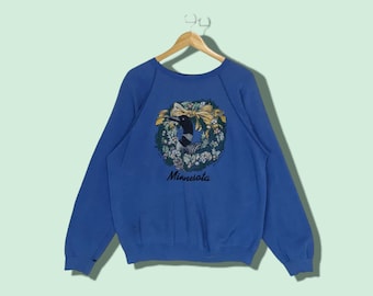 Puede incluir: Una sudadera azul con un diseño de corona colorido que presenta un pájaro y la palabra "Minnesota".