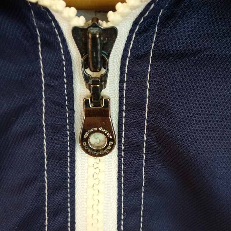 May include: Close-up of a zipper on a navy blue jacket with white stitching. The zipper pull has the words "Sport Surplus" and "Coupes" engraved on it.