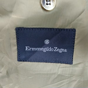 May include: A close-up of a dark blue label with white stitching around the edges. The label says "Ermenegildo Zegna" in white text with a small circle logo above the text.