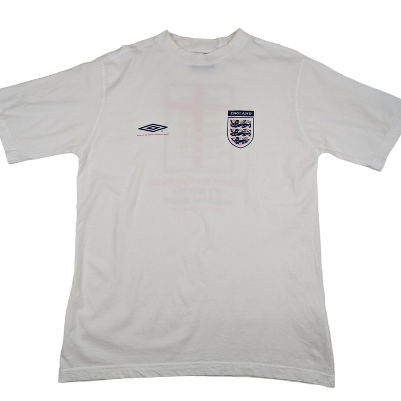 May include: White t-shirt with a red and blue England crest and a small, black and white logo of a diamond shape with the text "umbro" above it.