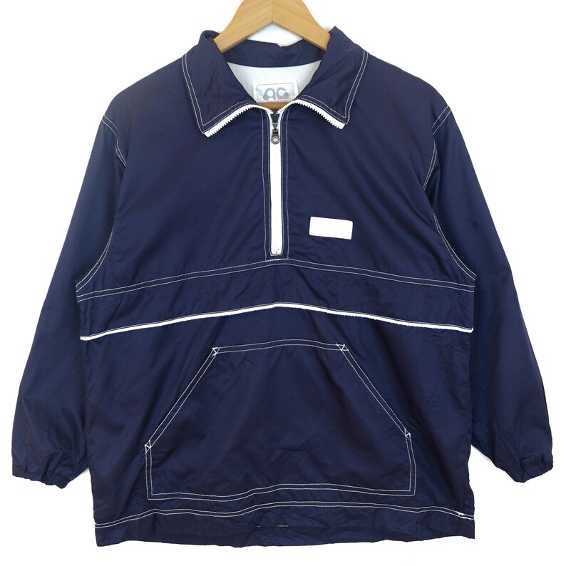 May include: A navy blue windbreaker jacket with white stitching and a zipper closure. The jacket has a large pocket on the front and a small white label with the letters "OC" on the collar.