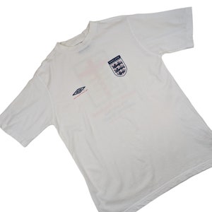May include: White short-sleeved t-shirt with a red cross and the England football team crest. The shirt has a blue and white diamond logo on the left side and the text "ENGLAND" above the crest.