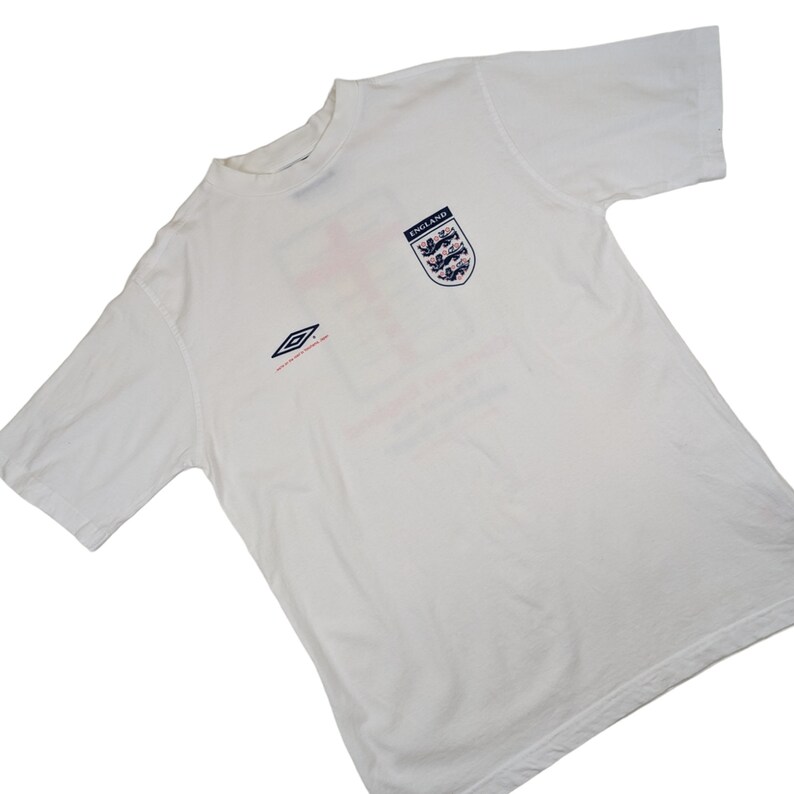 May include: White short-sleeved t-shirt with a red cross and the England football team crest. The shirt has a blue and white diamond logo on the left side and the text "ENGLAND" above the crest.