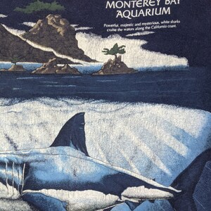 Peut inclure: Un t-shirt bleu avec un graphique d'un grand requin blanc nageant dans l'oc&eacute;an. Le texte "Monterey Bay Aquarium" est imprim&eacute; sur le t-shirt. Le texte "Powerful, majestic and mysterious, white sharks cruise the waters along the California coast." est imprim&eacute; sous le nom de l'aquarium.