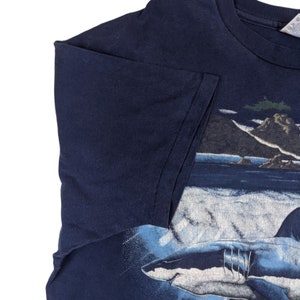 Peut inclure: Un t-shirt bleu marine avec un imprim&eacute; graphique d'un grand requin blanc nageant dans l'oc&eacute;an. Le requin est blanc avec des marques grises et est entour&eacute; d'eau bleue et de vagues blanches. L'arri&egrave;re-plan du t-shirt est de couleur bleu fonc&eacute;.