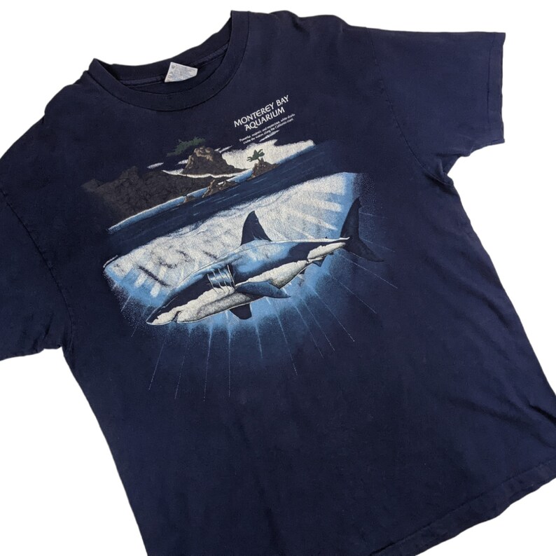 Peut inclure: Un t-shirt bleu marine avec un motif repr&eacute;sentant un grand requin blanc nageant dans l'oc&eacute;an. Le texte "Monterey Bay Aquarium" est imprim&eacute; sur le t-shirt.