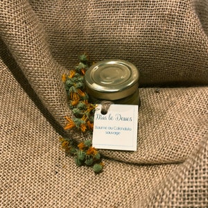 May include: A small glass jar with a gold lid, containing a balm, sits on a textured brown surface. A sprig of dried orange and green flowers is next to the jar. A small tag with text is attached to the jar.