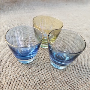 May include: Three small glass shot glasses. Two of the glasses are blue with a gold rim and one glass is yellow with a gold rim.
